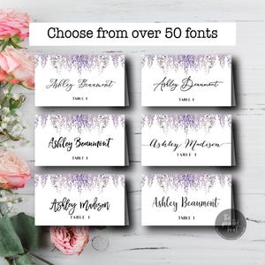 May include: White place cards with floral design and calligraphy names, with the text "Choose from over 50 fonts." The cards feature names like "Ashley Beaumont" and "Ashley Madison." Pink roses and greenery are in the background.