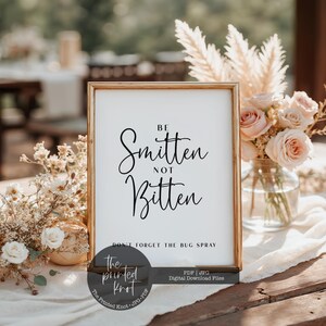 Op de afbeelding: Een ingelijst bord met de tekst "Be Smitten Not Bitten" in zwarte letters. Daaronder staat "Don't Forget The Bug Spray". Het bord staat op een tafel met bloemstukken, ideaal voor een bruiloft of een evenement in de open lucht.