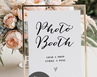 May include: A gold framed sign with the words "Photo Booth" in elegant black script. Below, it reads "Grab a prop, strike a pose" with a heart symbol. The sign is set against a backdrop of soft-colored flowers.