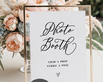 May include: A gold-framed sign with the words "Photo Booth" in elegant script. Below, it reads "Grab a prop, strike a pose" with a heart symbol. The sign is set against a backdrop of flowers and greenery, suggesting a wedding or event.