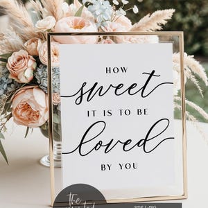 May include: A gold-framed sign with the words "How sweet it is to be loved by you" in black script. The sign is a digital download, and a bouquet of flowers is in the background. The sign is a digital download.
