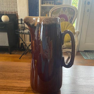 May include: A tall, brown ceramic pitcher with a handle and a spout. The pitcher has a glossy finish and a two-tone design, with a lighter brown rim. The pitcher is sitting on a wooden surface.
