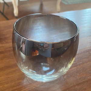 May include: A clear glass bowl with a silver-colored rim. The bowl has a rounded shape and sits on a wooden surface. The glass reflects the surrounding environment, creating a mirrored effect.