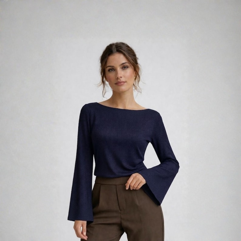 May include: A navy blue top with a boat neck and flared sleeves. The top is paired with brown trousers. The woman is standing in front of a white background.