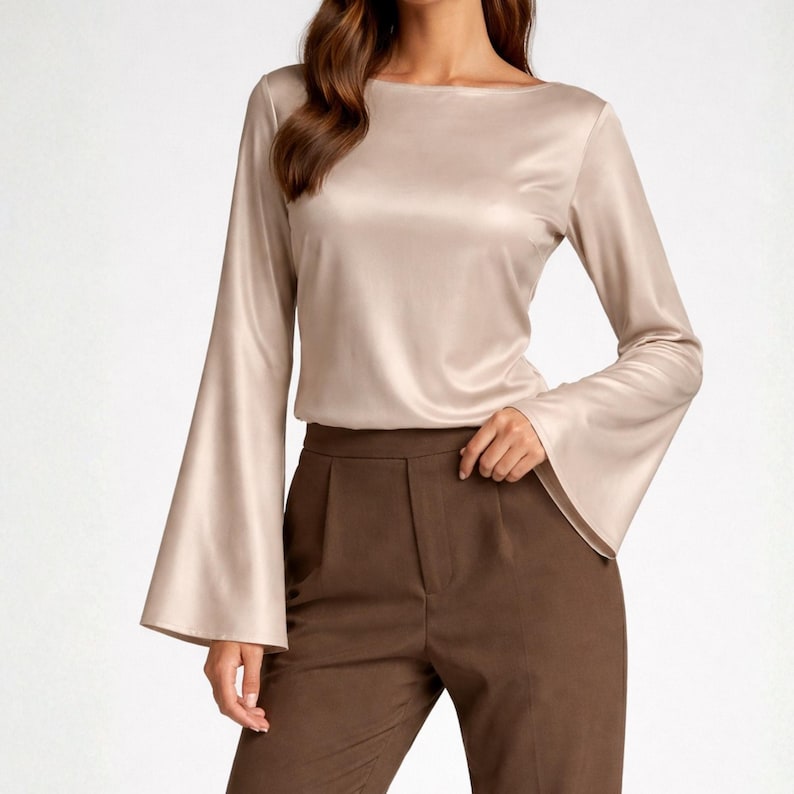 May include: A beige satin blouse with long, flared sleeves and a boat neckline. The blouse is paired with brown trousers. The fabric has a subtle sheen, and the overall look is elegant and sophisticated.