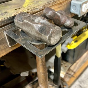 May include: Two metal hammers with wooden handles are hanging on a metal wall mount. The hammers are held in place by the metal mount, which is attached to a wooden surface.