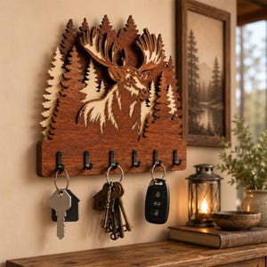 May include: A wall-mounted wooden key holder featuring a layered cutout design of a moose head and pine trees. The dark brown wood piece has six black metal hooks along the bottom edge, with three sets of keys hanging from the hooks on the left.