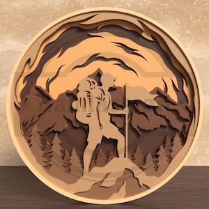 Puede incluir: Una obra de arte de madera en capas que representa a un excursionista con una mochila y una bandera en la cima de una montaña. La obra presenta un marco circular con una cordillera y un bosque en tonos marrones y beige. El excursionista está en silueta.