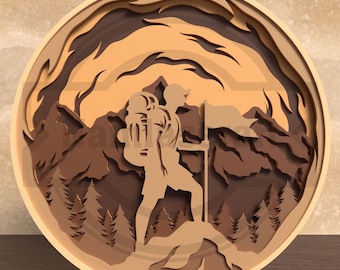 Puede incluir: Una obra de arte de madera en capas que representa a un excursionista con una mochila y una bandera en la cima de una montaña. La obra presenta un marco circular con una cordillera y un bosque en tonos marrones y beige. El excursionista está en silueta.