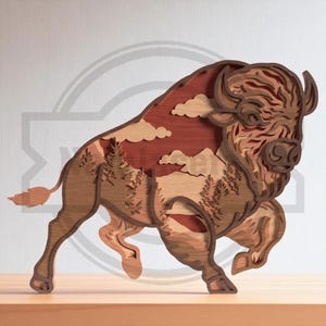 May include: A layered wooden bison sculpture with a landscape scene inside. The bison is brown with detailed carving. The landscape features trees, mountains, clouds, and a red sky. The sculpture is displayed on a wooden surface.