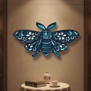 May include: A layered, three-dimensional wall art piece in the shape of a bee. The bee is primarily shades of blue, with intricate cut-out details in the wings and body. The artwork is mounted on a neutral-colored wall.