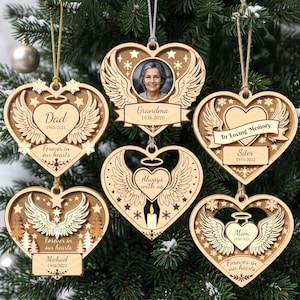 Op de afbeelding: Zes hartvormige houten ornamenten met engelvleugels, sterren en sneeuwvlokken. Ze bevatten teksten zoals Dad 1945-2021, Grandma 1938-2020, In Loving Memory Siter 1975-2022, Forever in our hearts Michael 1960-2023, Always with us en Mom 1950-2022.