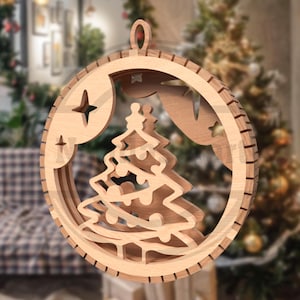 以下が含まれることがあります： 木製のクリスマスオーナメント。重ねられたデザインで、中央にクリスマスツリーがあり、星の切り抜きがある円形のフレームで囲まれています。明るい色の木材で作られており、吊り下げ用のループが付いています。