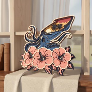Può includere: Un'opera d'arte in legno a strati raffigurante un polpo blu con fiori di ibisco rosa. Il design del polpo include una scena di tramonto con palme. L'opera d'arte è esposta su una superficie di legno vicino a una finestra.