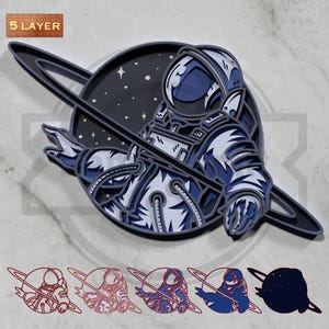 May include: A layered wooden art piece featuring an astronaut in a spacesuit against a backdrop of a planet with rings and stars. The astronaut is primarily blue and white. The words "5 LAYER" are in the upper left corner.