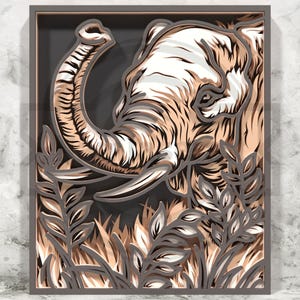 May include: A multi-layered wooden artwork depicting an elephant's head and trunk, with detailed fur and tusks. The piece uses shades of brown, white, and grey, with foliage in the foreground. The artwork is framed in a grey border.
