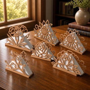 Könnte beinhalten: Eine Sammlung von sechs hellbraunen Serviettenhaltern aus Holz mit filigranen, lasergeschnittenen Blumenmotiven wie Sonnenblumen, Rosen und Kirschblüten, die weiße, dreieckig gefaltete Servietten auf einer dunklen Holzoberfläche halten.
