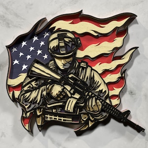 Puede incluir: Una obra de arte de madera en capas que representa a un soldado sosteniendo un rifle, sobre un fondo de la bandera estadounidense. La bandera presenta franjas rojas y blancas con un campo azul de estrellas. El soldado viste un uniforme de combate, con casco y chaleco antibalas.