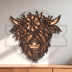 Puede incluir: Arte de pared de madera que representa la cabeza de una vaca de las Tierras Altas con una corona floral. La obra de arte est&aacute; hecha de madera en capas, mostrando un dise&ntilde;o detallado con diferentes tonos de marr&oacute;n. La vaca tiene pelo largo y cuernos curvados, y las flores est&aacute;n intrincadamente talladas.