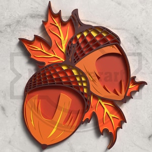 Puede incluir: Una obra de arte en papel en capas que presenta dos bellotas y hojas de otoño en tonos naranja, amarillo y marrón. El diseño tiene un efecto 3D, con intrincados detalles de corte que crean profundidad y sombra. La obra de arte está sobre un fondo gris claro.