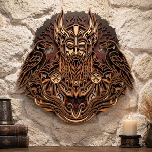 May include: A multi-layered wooden wall art piece featuring a central bearded figure wearing a winged helmet, flanked by two ravens. The design uses various shades of brown and tan wood, creating depth through intricate cutouts and stacked layers.