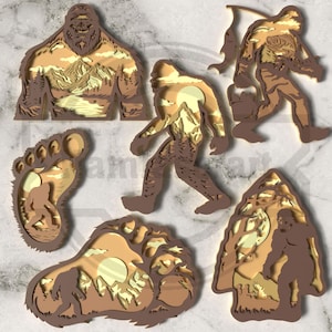 May include: A selection of layered wooden Bigfoot-themed art pieces. The pieces feature various depictions of Bigfoot in a brown and tan colour palette, with mountain and forest scenery. The collection includes Bigfoot figures, footprints, and a mountain scene.
