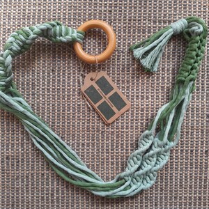 Puede incluir: Un macetero de macramé verde y azul con un anillo de madera. El macetero tiene un borla en cada extremo.