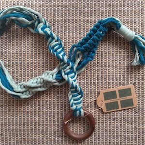 Könnte beinhalten: Ein Makramee-Pflanzenhänger mit einem Holzring, hergestellt aus blauem und grünem Baumwollschnur. Der Hänger hat ein Etikett mit der Aufschrift "Handmade in Etsy".