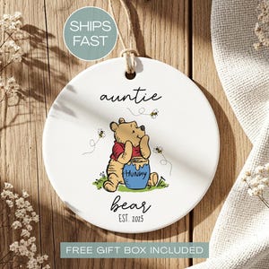 May include: A white ceramic ornament with a Winnie the Pooh design. The ornament features the words "auntie bear" and "EST. 2025". The image includes a cartoon bear, honey pot, and bees. A string is attached for hanging.