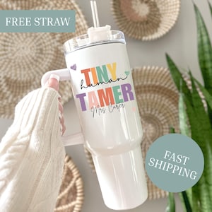 May include: White tumbler with a handle and a clear lid, featuring the text "TINY human TAMER Mrs Carter" in colourful letters. Includes a clear straw. The tumbler is held by a person wearing a cream-coloured sweater.