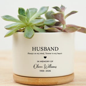May include: A white ceramic planter with a succulent plant, inscribed with "HUSBAND" and "In Memory of Oliver Williams 1956-2026". The planter rests on a wooden base, ideal for home decoration.