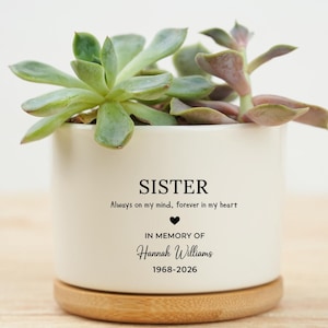 Peut inclure: Un pot de fleurs en céramique blanc avec une plante succulente, posé sur une base en bois. Le pot porte l'inscription "SISTER", la phrase "Always on my mind, forever in my heart" et "In memory of Hannah Williams 1968-2026".