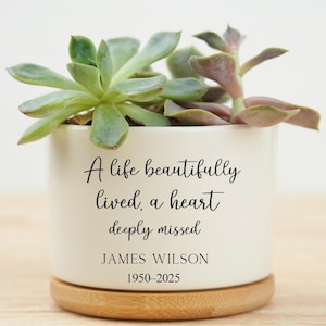 May include: A white ceramic planter with a succulent plant. The planter has the inscription "A life beautifully lived, a heart deeply missed" and the name "JAMES WILSON 1950-2025". The planter sits on a wooden base.