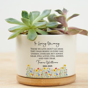 Op de afbeelding: Een witte keramische plantenpot met een houten basis, met een vetplant. De pot heeft de tekst "In Loving Memory" en een herdenkingsboodschap, samen met de naam Thomas Williams en de jaren 1959-2025. Bloemenontwerp aan de onderkant.