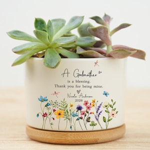 Puede incluir: Una maceta de cerámica blanca con una planta suculenta. La maceta tiene un diseño floral y el texto "A Godmother is a blessing. Thank you for being mine." Se apoya en una base de madera.