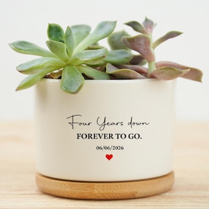May include: A white ceramic planter with a wooden base, holding a succulent plant. The planter has the text "Four Years down FOREVER TO GO." with a heart and the date 06/06/2026. The succulent has green and purple leaves.