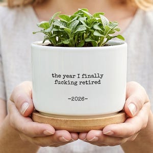 May include: A white ceramic cylindrical plant pot with a bamboo saucer base. The pot features black typewriter-style text that reads: the year I finally fucking retired -2026-. A small green leafy plant with white veined foliage grows inside.