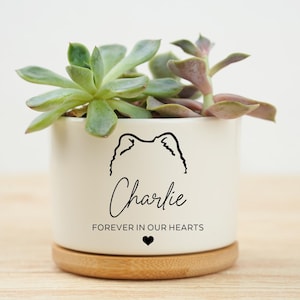 Könnte beinhalten: Ein weißer Keramik-Pflanztopf mit einer Sukkulentenpflanze. Der Topf hat eine schwarze Strichzeichnung eines Hundegesichts, den Namen "Charlie" und den Text "FOREVER IN OUR HEARTS" mit einem Herzsymbol. Er steht auf einem runden Holzsockel.