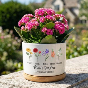May include: A white ceramic plant pot with a wooden base, filled with bright pink flowers. The pot features floral illustrations and the text "Mom's Garden, Love Grows Here."