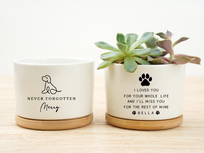 May include: Two white ceramic planters with bamboo bases. One features a dog outline and the text "NEVER FORGOTTEN Merry". The other has a paw print and the text "I LOVED YOU...BELLA". One planter holds a succulent.