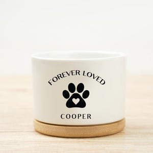 May include: A white ceramic planter with the words "FOREVER LOVED" above a black paw print with a heart, and the name "COOPER" below. The planter sits on a round, light brown wooden base.