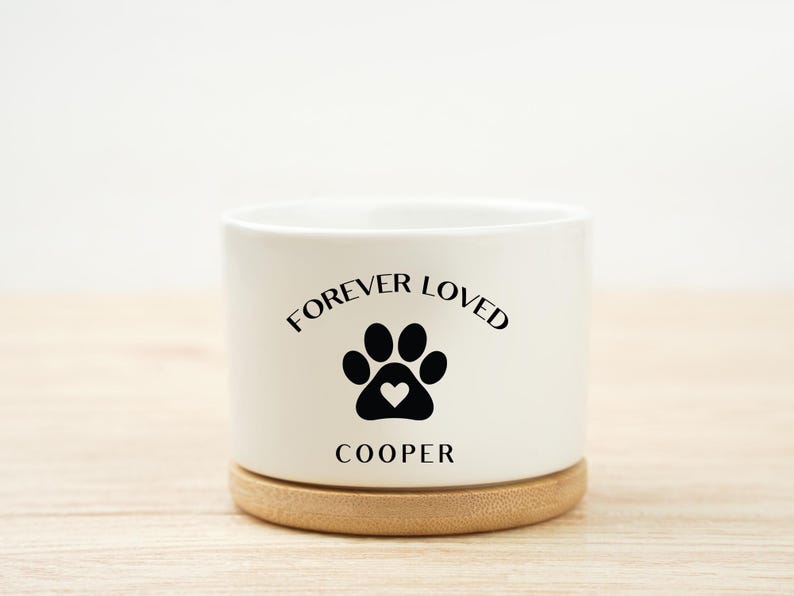 May include: A white ceramic planter with the words "FOREVER LOVED" above a black paw print with a heart, and the name "COOPER" below. The planter sits on a round, light brown wooden base.