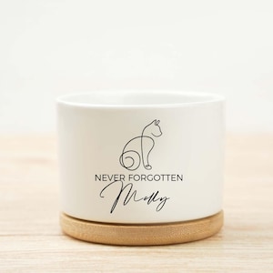 May include: A white ceramic planter with a bamboo base. The planter features a black line drawing of a cat and the words "NEVER FORGOTTEN" and "Molly" in a script font. A memorial item for a pet.