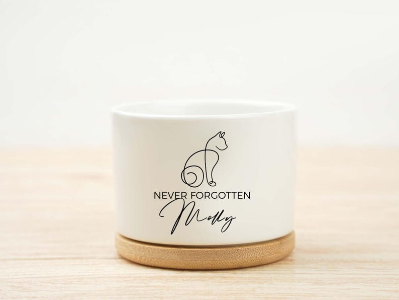 May include: A white ceramic planter with a bamboo base. The planter features a black line drawing of a cat and the words "NEVER FORGOTTEN" and "Molly" in a script font. A memorial item for a pet.