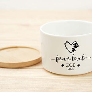May include: A white ceramic pet bowl with a wooden coaster. The bowl features a black heart with paw prints, the words "forever loved", the name "ZOE", and the year "2025".