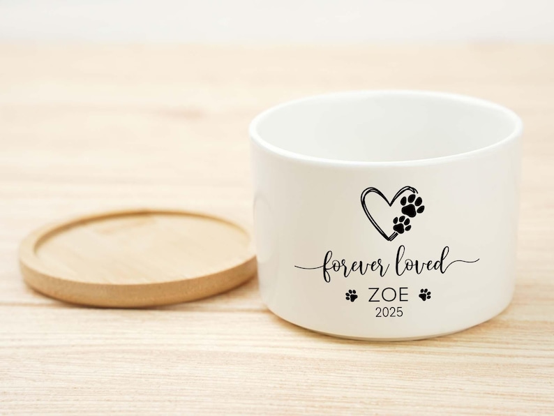 May include: A white ceramic pet bowl with a wooden coaster. The bowl features a black heart with paw prints, the words "forever loved", the name "ZOE", and the year "2025".