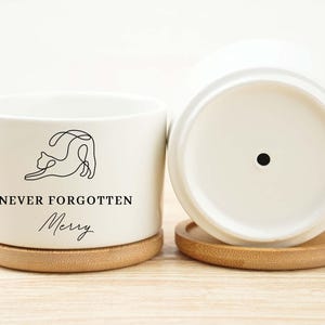 May include: White ceramic planter with a minimalist cat illustration and the text "NEVER FORGOTTEN Merry". The planter sits on a round bamboo coaster. Another planter is visible, upside down.