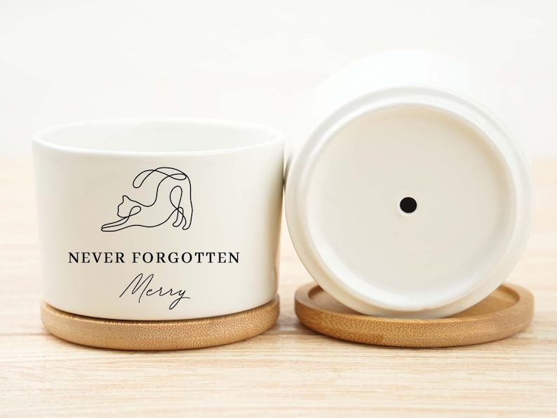 May include: White ceramic planter with a minimalist cat illustration and the text "NEVER FORGOTTEN Merry". The planter sits on a round bamboo coaster. Another planter is visible, upside down.