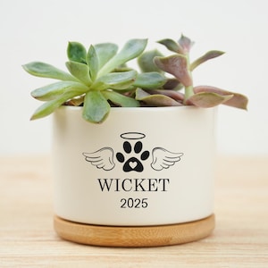 May include: A white ceramic planter with a succulent plant. The planter has a paw print design with angel wings and the name "WICKET" and the year "2025" in black. The planter sits on a wooden base.