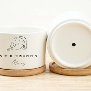 May include: White ceramic planter with a minimalist cat illustration and the text "NEVER FORGOTTEN Merry". The planter sits on a round bamboo coaster. Another planter is visible, upside down, showing the drainage hole.